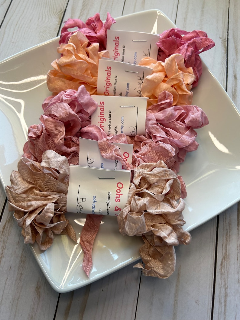May include: A collection of ruffled silk ribbons in shades of pink, peach, and beige, displayed on a white square plate. Each ribbon is bundled and tagged with a small white label. The ribbons are arranged on a light wood surface.