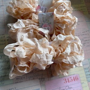 May include: A close-up of a bundle of ivory-colored ruffled ribbon. The ribbon is tied together with a piece of twine. There are three small paper tags with handwritten text on them. The text on the tags reads "Croq Oat", "Sarah Broste", and "Liam Olsen Free Your Heart".