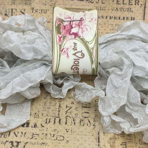 May include: A spool of silver ruffled trim with a vintage label that reads "Aux Violets" in French. The label features a floral design with pink flowers and green leaves.