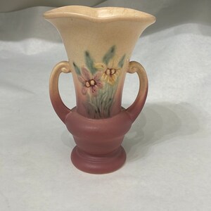 May include: A small, cream-colored ceramic vase with two handles. The vase has a floral design with pink and yellow flowers.