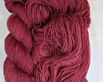 Puede incluir: Primer plano de hilo de color burdeos, presentado en madeja y ovillo. Se aprecian la textura y las variaciones de color. El hilo parece suave, ideal para proyectos de punto o ganchillo.