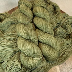 May include: Close-up of several skeins of olive green yarn. The yarn is tightly wound into neat bundles, with loose strands spread around. The color is a muted, earthy green, perfect for knitting or crocheting projects.