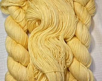 Puede incluir: Varios ovillos de hilo amarillo pálido están dispuestos verticalmente. El hilo está enrollado en madejas apretadas, con hebras sueltas visibles. El color es un amarillo suave y mantecoso, lo que sugiere una fibra natural. Ideal para proyectos de punto y ganchillo.