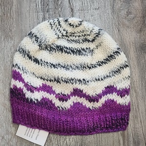 May include: A purple, white, and black striped knitted hat with a wavy pattern. The hat is made of wool and has a soft, comfortable feel.