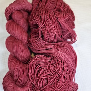 May include: Two skeins of burgundy-colored yarn. One skein is tightly wound, while the other is loose and appears ready for use. The yarn has a soft, textured appearance, suitable for knitting or crocheting projects.