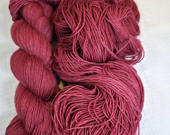 Puede incluir: Dos madejas de hilo de color burdeos. Una madeja está bien enrollada, mientras que la otra está suelta y parece lista para usar. El hilo tiene un aspecto suave y texturizado, adecuado para proyectos de tejido o ganchillo.