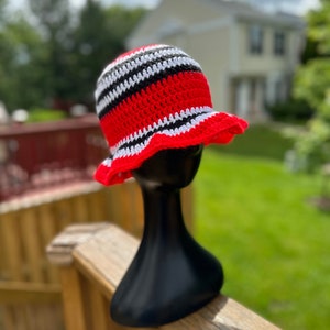 Op de afbeelding: Een rode, witte en zwarte gestreepte crochet bucket hat met een brede rand. De hoed wordt getoond op een zwart mannequinhoofd.
