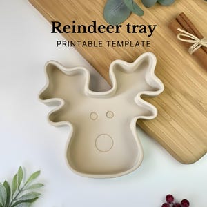 Op de afbeelding: Een beige rendiervormige schaal met de tekst "Reindeer tray PRINTABLE TEMPLATE". De schaal heeft een cirkelvormige neus en twee kleine cirkelvormige ogen. De schaal ligt op een houten snijplank.