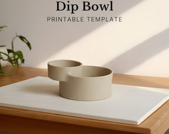 May include: A beige ceramic dip bowl with a circular main bowl and a smaller attached bowl. The text "Dip Bowl PRINTABLE TEMPLATE" is displayed above the bowl. The bowl is on a white rectangular surface, placed on a wooden table.