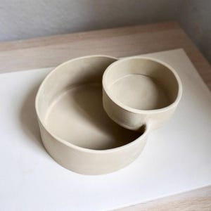 Może przedstawiać: Beżowa ceramiczna miska do serwowania o okrągłym kształcie. Naczynie ma większą, otwartą miskę połączoną z mniejszą, okrągłą miską. Miski są gładkie i mają matowe wykończenie, odpowiednie do serwowania przekąsek lub dip&oacute;w.