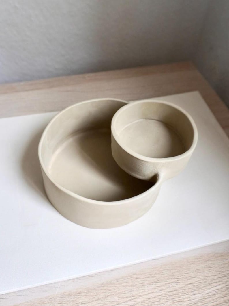 Może przedstawiać: Beżowa ceramiczna miska do serwowania o okrągłym kształcie. Naczynie ma większą, otwartą miskę połączoną z mniejszą, okrągłą miską. Miski są gładkie i mają matowe wykończenie, odpowiednie do serwowania przekąsek lub dip&oacute;w.