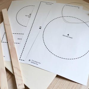 Może przedstawiać: Zestaw materiał&oacute;w rzemieślniczych, w tym białe szablony papierowe z przerywanymi liniami i tekstem, drewniane listwy i ż&oacute;łty przedmiot. Szablony papierowe zawierają tekst "Dip bowl base" i "Cut down the dotted line".