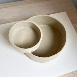 Może przedstawiać: Para beżowych ceramicznych misek, jedna mniejsza i połączona z większą. Miski mają gładkie, matowe wykończenie i są ustawione na białym i jasnym drewnianym tle. Projekt jest prosty i nowoczesny.