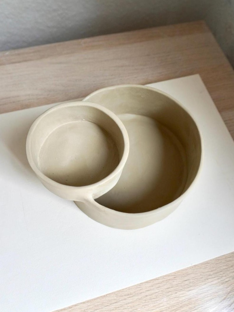 Może przedstawiać: Para beżowych ceramicznych misek, jedna mniejsza i połączona z większą. Miski mają gładkie, matowe wykończenie i są ustawione na białym i jasnym drewnianym tle. Projekt jest prosty i nowoczesny.