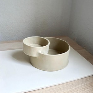 Może przedstawiać: Beżowa ceramiczna miska z mniejszą, dołączoną miską. Miski są okrągłe, z gładkim, matowym wykończeniem. Projekt jest minimalistyczny i nowoczesny, odpowiedni do serwowania lub ekspozycji.