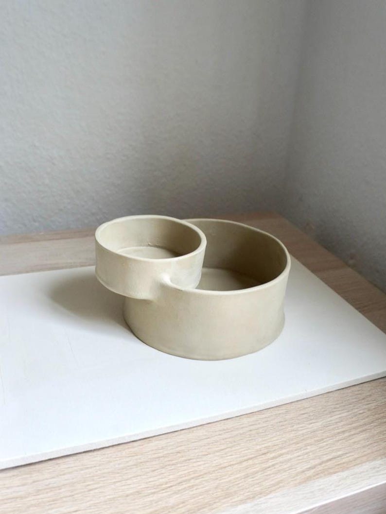 Może przedstawiać: Beżowa ceramiczna miska z mniejszą, dołączoną miską. Miski są okrągłe, z gładkim, matowym wykończeniem. Projekt jest minimalistyczny i nowoczesny, odpowiedni do serwowania lub ekspozycji.