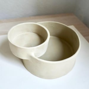 Może przedstawiać: Beżowa ceramiczna miska do serwowania z małą, dołączoną miseczką. Naczynie ma okrągłą podstawę z mniejszą, okrągłą miseczką przymocowaną z boku. Projekt jest minimalistyczny i nowoczesny, z matowym wykończeniem.
