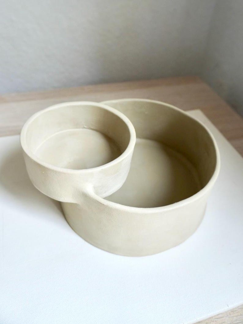 Może przedstawiać: Beżowa ceramiczna miska do serwowania z małą, dołączoną miseczką. Naczynie ma okrągłą podstawę z mniejszą, okrągłą miseczką przymocowaną z boku. Projekt jest minimalistyczny i nowoczesny, z matowym wykończeniem.
