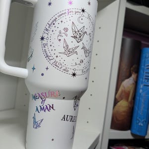 May include: White insulated travel mug with a handle and a holographic lid. The mug features a design with origami cranes, stars, and the text "I'll always come for you." Additional text includes "Measure of Aman" and "Aurel."