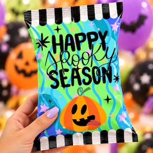 May include: A rectangular snack bag with black and white striped borders at the top and bottom. The center features a bright green and blue wavy background with pink stars and black text that reads HAPPY SPOOKY SEASON above a cartoon pumpkin.