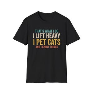 Könnte beinhalten: Schwarzes T-Shirt mit dem Text "THAT'S WHAT I DO I LIFT HEAVY I PET CATS AND I KNOW THINGS" in Türkis, Weiß, Gold und Rot. Das Shirt hat einen Rundhalsausschnitt und kurze Ärmel.