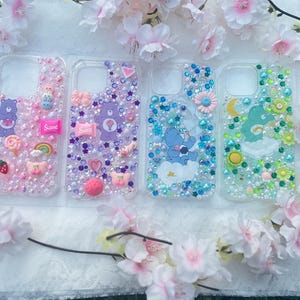 May include: Four clear phone cases decorated with Care Bears, bows, and rhinestones. One case has the word "Sweet" on it. The cases are surrounded by pink and white flowers.