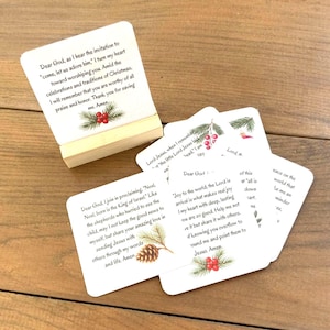 May include: A set of square white prayer cards featuring printed religious text and illustrations of pine branches with red berries or pinecones. One card is displayed upright on a small wooden stand, while others are fanned out underneath.