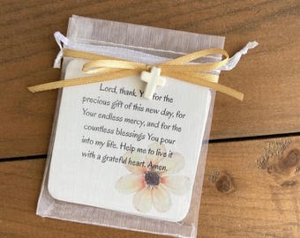 May include: A white square card inside a sheer organza drawstring bag, tied with a gold ribbon and a small white cross charm. The card features a printed prayer starting with Lord, thank You for the precious gift of this new day, and a flower.