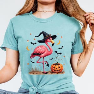 May include: A teal short-sleeve t-shirt featuring a graphic of a pink flamingo wearing a black witch hat. The flamingo stands near a carved orange jack-o'-lantern pumpkin. The design includes small black bats, orange stars, and yellow crescent moons.