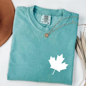 May include: A teal Comfort Colors t-shirt with a white maple leaf graphic. The shirt has a crew neck and short sleeves. A gold necklace with a pendant is draped across the shirt. The shirt is size small.