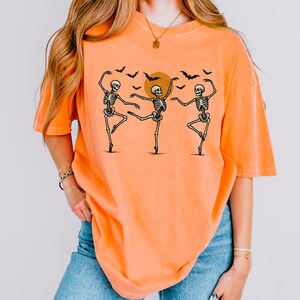 May include: An orange short-sleeve t-shirt featuring a graphic print of three black skeletons dancing in ballet poses. Above the skeletons is a solid orange circle representing a moon, surrounded by several small black flying bat silhouettes.