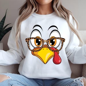 Può includere: Felpa bianca con un volto di tacchino dei cartoni animati con gli occhiali. Il tacchino ha grandi occhi, un becco giallo e un bargiglio rosso. Gli occhiali hanno un motivo leopardato. La felpa è un capo casual e comodo.