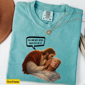 May include: Sea foam green t-shirt with a graphic of Jesus whispering to a man, with the text "YOU ARE NOT JESUS, SNAP OUT OF IT" in a speech bubble. The shirt has a Comfort Colors tag and a small necklace.