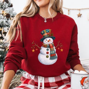 May include: A dark red long-sleeve sweatshirt featuring a central graphic of a snowman wearing a colorful patterned top hat and scarf. The snowman has three buttons on its torso and holds two thin branches with hanging yellow star ornaments.
