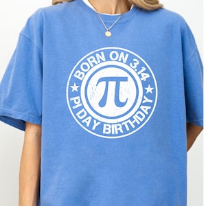 Puede incluir: Una camiseta azul claro con un gráfico circular blanco que dice "BORN ON 3.14 PI DAY BIRTHDAY" alrededor de un símbolo Pi. La camiseta tiene mangas cortas y cuello redondo.