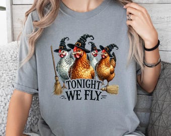 May include: A grey short-sleeved t-shirt featuring a graphic of four chickens wearing black witch hats. The chickens are positioned above the text "TONIGHT WE FLY" and two straw broomsticks are illustrated on either side of the text.