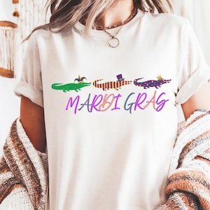 Puede incluir: Camiseta color crema con tres caimanes de dibujos animados, cada uno con un sombrero diferente. El texto "MARDI GRAS" está escrito en cursiva morada y verde debajo. La camiseta tiene mangas cortas.