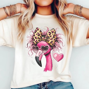 May include: A cream-colored t-shirt featuring a graphic of a pink flamingo with messy hair, wearing heart-shaped sunglasses and a large leopard print bow. A solid pink heart is positioned to the right of the flamingo head illustration.