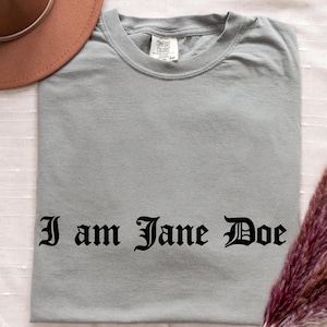 May include: A grey short-sleeved t-shirt with a crew neckline and the text "I am Jane Doe" printed in black gothic-style lettering across the chest. The shirt features a white interior neck label with the brand name Comfort Colors visible.