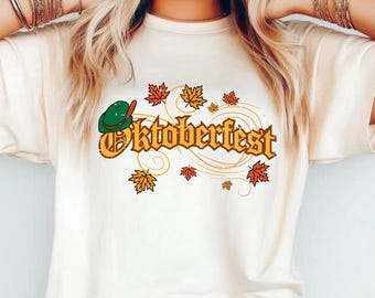 May include: A cream-colored t-shirt featuring a graphic print with the text "Oktoberfest" in a bold, orange-brown font. The design includes a green alpine hat with a red feather and several scattered orange and yellow maple leaves.