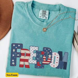 May include: Sea foam green Comfort Colors t-shirt with the word "FREEDOM" appliquéd across the front in red, white, and blue patriotic patterns. A gold necklace with a pendant rests on the shirt. A brown hat is partially visible.