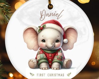 May include: A circular Christmas ornament with a baby elephant wearing a Santa hat and a red and green sweater. The ornament is inscribed with the name "Daniel" and the text "FIRST CHRISTMAS".