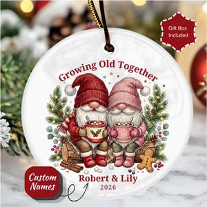May include: A round white ceramic ornament featuring two gnomes in red and pink hats sitting on a log, holding mugs with the text Growing Old Together, Robert & Lily, and 2026. The design includes pine branches, berries, and a gingerbread man.