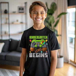 May include: Black t-shirt with a pixelated video game controller design. The shirt displays the text "KINDERGARTEN LEVEL BEGINS" in green and white. The controller graphic incorporates green, brown, red, yellow, and blue colours.