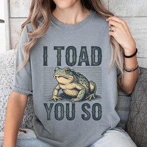 May include: Grey t-shirt featuring the phrase "I TOAD YOU SO" with a detailed illustration of a green and tan toad. The shirt is a solid colour and has short sleeves.