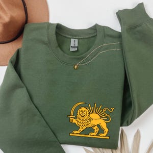 May include: An olive green sweatshirt with a gold lion and sun emblem. The design shows a lion holding a sword, with a sunburst behind it. A gold necklace with a pendant is also visible.