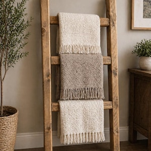 May include: A wooden blanket ladder displays three textured throws in cream and taupe. The throws have fringed edges and are draped over the ladder's rungs. The ladder is made of light brown wood and leans against a neutral-coloured wall.