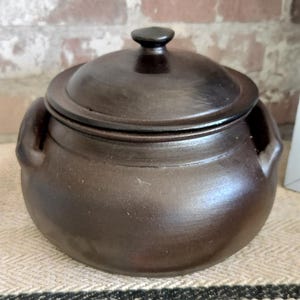 Può includere: Un vaso di ceramica marrone scuro con coperchio. Il vaso ha due manici e un piccolo pomello sulla parte superiore del coperchio. Il vaso è appoggiato su un tessuto a righe. Lo sfondo è un muro di mattoni.