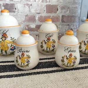 May include: Set of five ceramic kitchen canisters with lids. Each canister is off-white with an orange lid and trim. They feature a colorful illustration of a person in a yellow coat and the words "Sel", "Cafe", "Poivre", "Epices", and "Sucre".