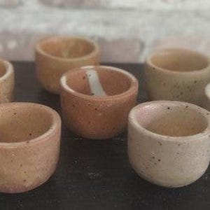 May include: Seven small, handmade ceramic cups in shades of tan and brown. The cups have a speckled texture and a rounded shape. Ideal for serving small portions, they are crafted from ceramic.